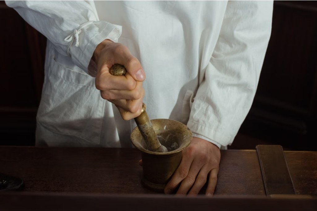 A pharmacist in a white coat grinding ingredients in a brass mortar and pestle